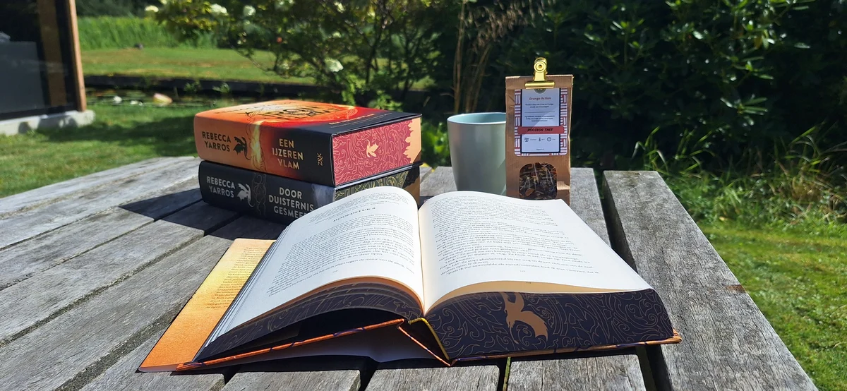 Een open boek, boeken van andere auteurs en een drankje op een houten tuintafel